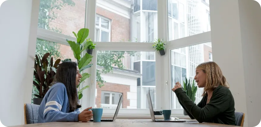 Two girls having a conversation, while sitting in front of each other with their laptops.